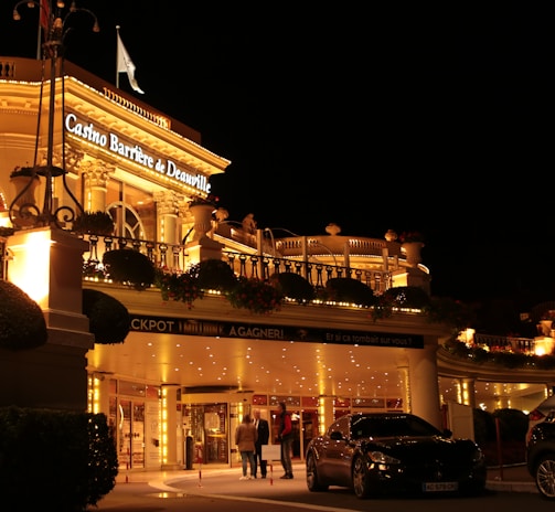 A luxurious casino at night, illuminated with warm, golden lights. The building features elegant architecture with columns and decorative elements. A few people are walking towards the entrance, and there are a couple of parked cars nearby.